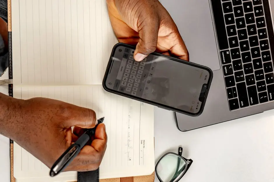 Člověk zapisuje odpracovaný čas do sešitu vedle telefonu a notebooku.
