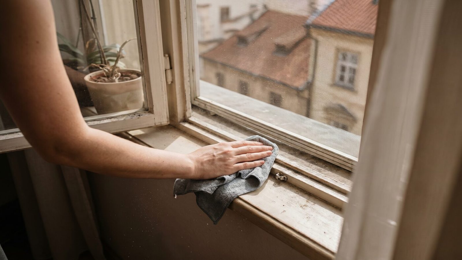 Detail parapetu a záclony v bytě, kde někdo utírá jemný prach vlhkou utěrkou z mikrovlákna.