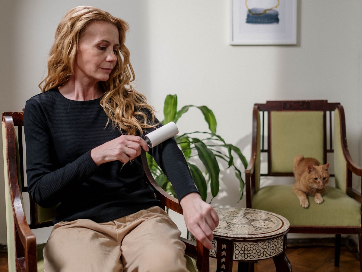 Odstraňování chlupů z textilu pomocí lepivého válečku, zatímco poblíž sedí kočka.